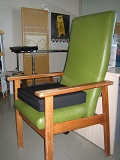 Image shows a chair with arms and a firm, level seat with a cushion to raise the seated surface.  Cushion attaches to the chair for safety. 
