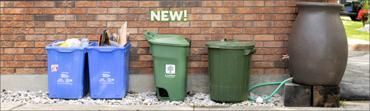 Photograph of two blue recycling bins, the new green bin, a trash bin, and a rain barrel at the side of a house.