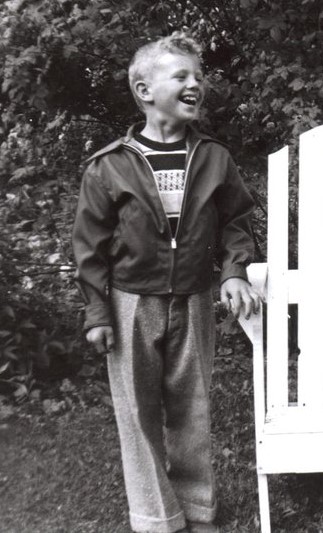 A black and white photo of Dave Gast when he was eight-years-old in 1952. The little boy is standing and smiling.