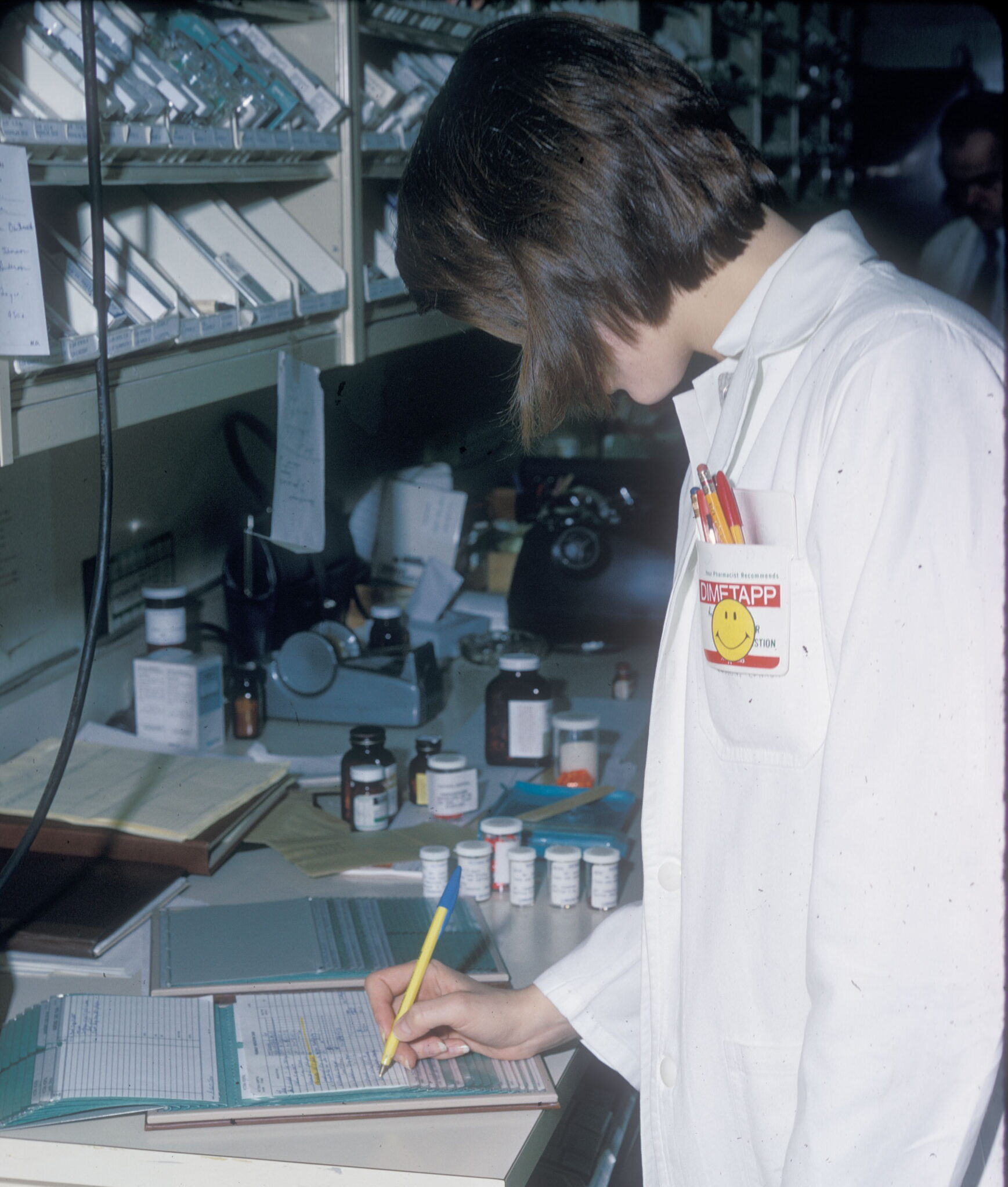 A pharmacist in 1972 recording a dispensed prescription onto a patient profile, kept in a Kardex folder. A typewriter was used to type up the label for the prescription bottle. A copy may have also been placed on the patient profile in the Kardex folder.