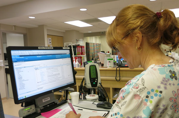 Female nurse in hospital scrubs looking at a computer screen at a nurse station.