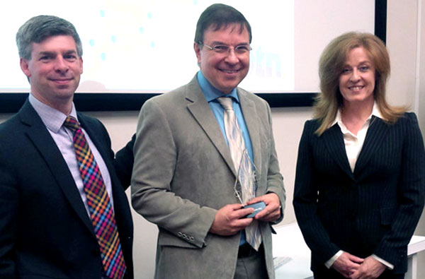 Two men in suits and ties standing with a woman in a suit in front of a board room, smiling.