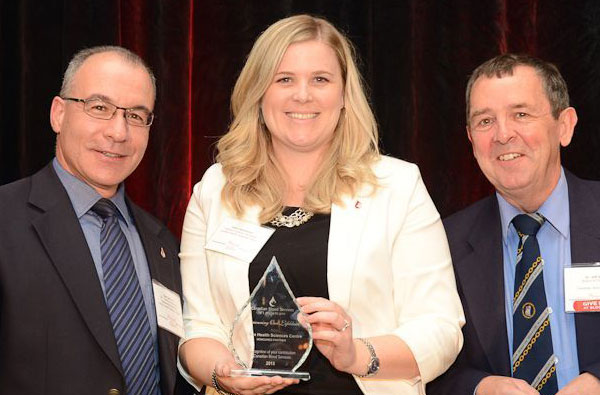 Two men in suits standing on either side of a female with blonde hair accepting an award that is made of glass and shaped like a rain drop.