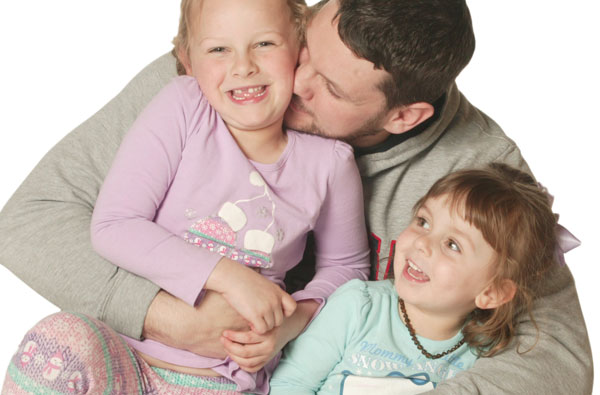 Father sitting on the ground with two young girls sitting on his lap with his arms around them smiling