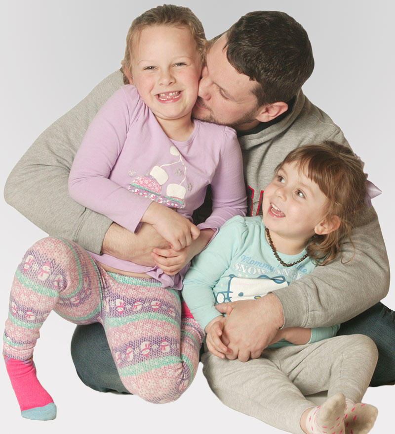 Father sitting on the ground with his arms around his two young daughters, kissing one of them on the cheek.