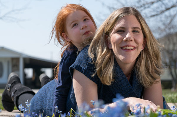 Young mother laying on the grass with her young daughter sitting on her back both smiling at the camera