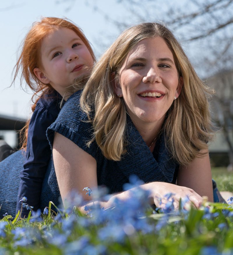 Mother laying on the grass with her young daughter laying on her back, both smiling at the camera. 