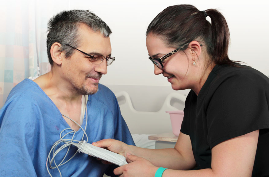 Female nurse with dark hair, glasses, and black shirt, holding a testing device up to a man with glasses in a blue hospital gown.