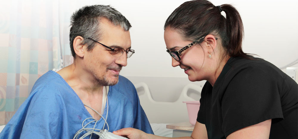 Female nurse with dark hair, glasses, and black shirt, holding a testing device up to a man with glasses in a blue hospital gown.
