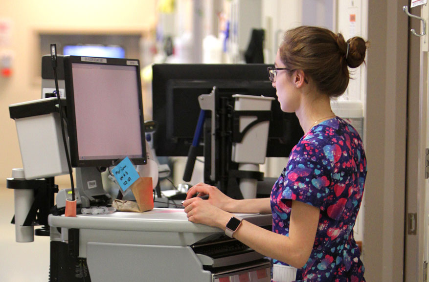 Female nurse wearing glasses and purple scrubs working on a computer at a nurse station.