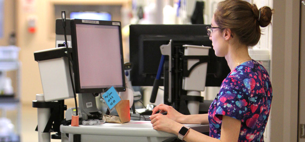 YFemale nurse wearing glasses and purple scrubs working on a computer at a nurse station.