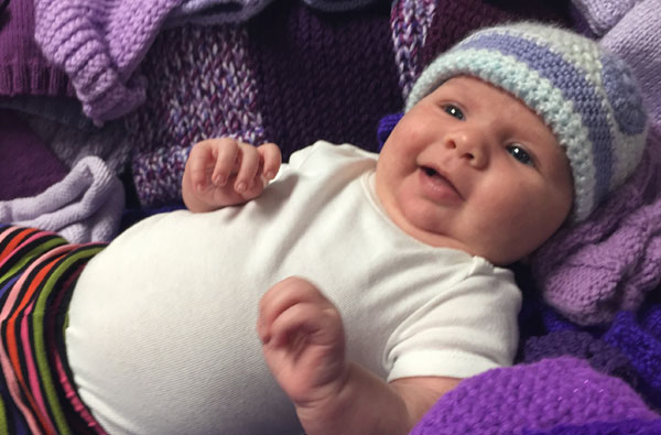 Newborn baby in a white shirt, striped pants, and knitted hat, laying among purple knitted caps.