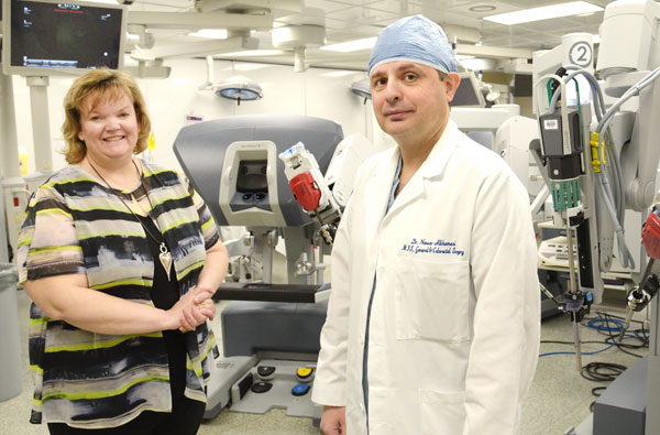 Male doctor in white doctor coat and blue head mask standing beside a woman with short red hair in front of a piece of hospital equipment.