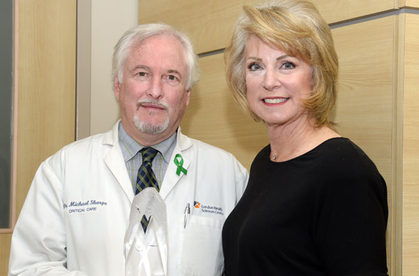 White haired man in a doctor’s white lab coat holding a small glass award standing beside a woman in a black dress shirt.