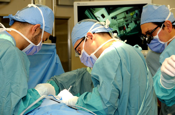 Three male surgeons in blue hospital gowns operating on a patient who is laying down on an operating table.