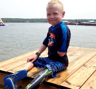 A young boy smiling at the camera while sitting on a dock in his swimsuit.