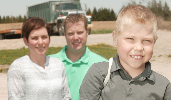 Mother and father behind their son standing outside in a field in front of a large truck.