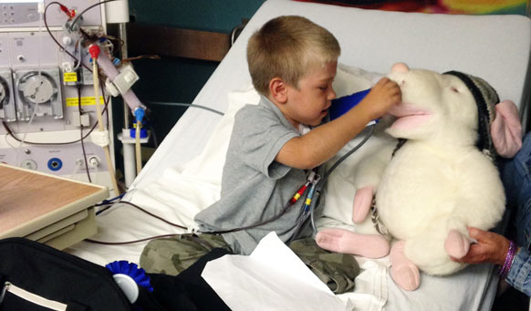 A young boy using medical instruments on stuffed animal while in hospital bed.