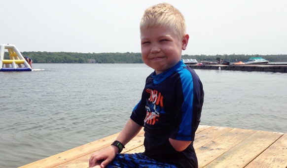 A young boy smiling at the camera while sitting on a dock in his swimsuit.