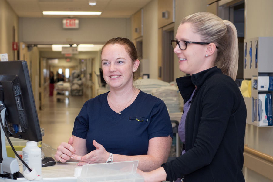 Two female nurses standing in a hospital hallway looking at a computer screen.