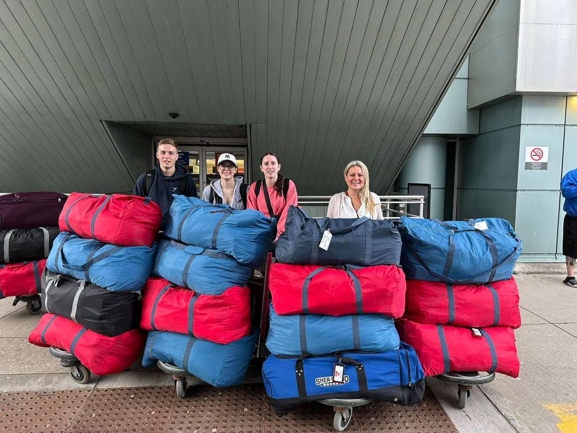 Four people stand behind three luggage carts stacked with large red and blue duffel bags. The scene is outside an airport.