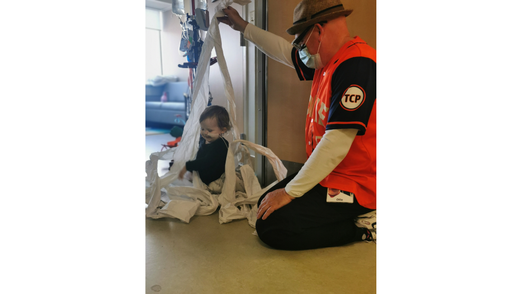 A therapeutic clown plays with a small child on the floor at a hospital. They are making a fort out of toilet paper.