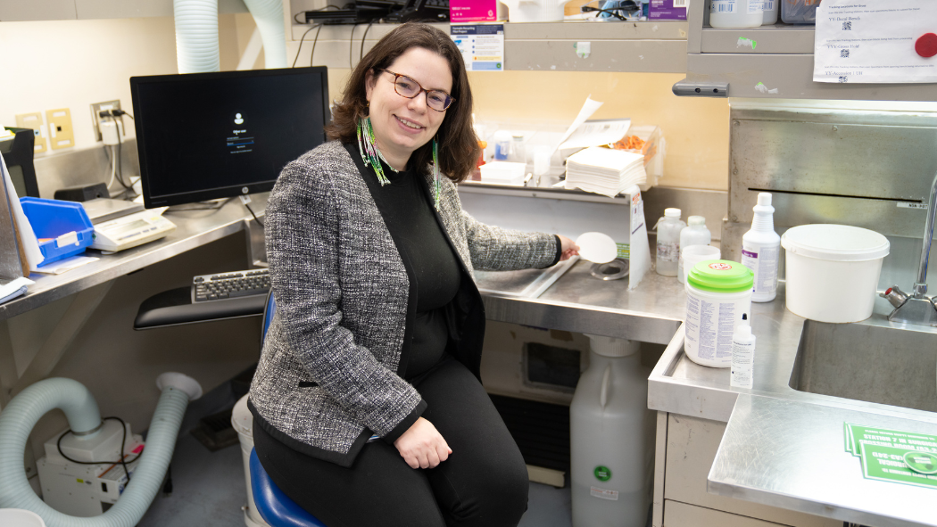 A person in a tweed jacket and glasses sits at a lab bench with a computer and various lab equipment.