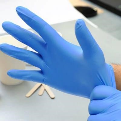 Close-up of a person putting on a blue disposable glove. The right hand is inside the glove while the left hand pulls it over the wrist. A white cup and wooden sticks are blurred in the background on a light surface.