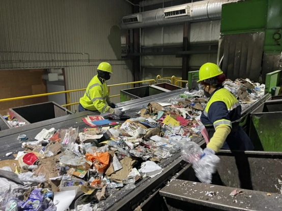 Photograph of line workers sorting through the incoming recyclable materials and pulling specific items, like plastic bags, into their own bin.