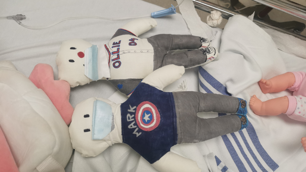Two handmade dolls sit on a hospital bed. One is named Mark, the other named Ollie.