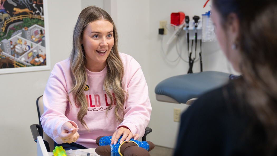 Person seated in a clinical exam room demonstrating an item on a doll while another person sits across from them, with medical equipment and wall artwork visible behind them.