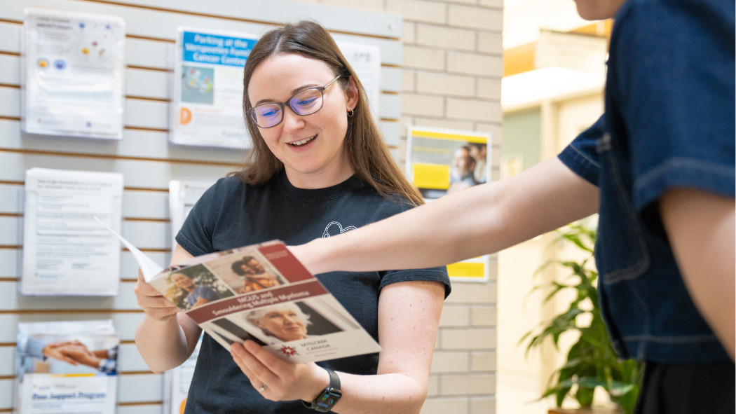 Person holding and reading a brochure while another person points to information on the page, with informational posters displayed on a wall behind them in a hospital setting.