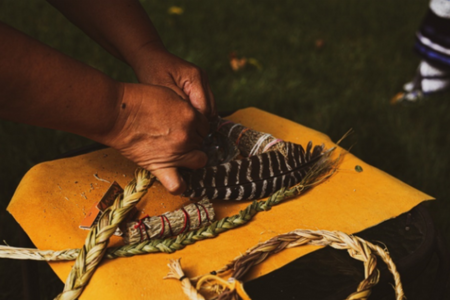 Image of hands working with sage, sweet grass, cedar, tobacco and a feather.