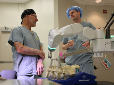 Dr. Timothy Carey and Dr. Patrick Thornley wearing blue scrubs and standing behind a Mazor robotic platform.