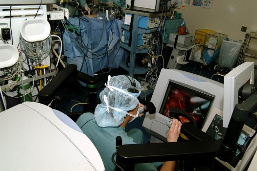 In the foreground you can see the patient and the three robotic arms working through three small incisions in the abdomen. Dr. Luke is seated at the robotic controls in the background moving the robotic arms to complete the surgery.