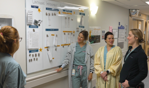 Four health-care professionals discuss information displayed on a whiteboard in a hospital corridor.