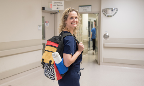 Smiling woman in scrubs stands in a hospital hallway with a colorful backpack over her shoulder, looking back at the camera.