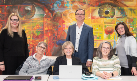 Seven adults pose for a group photo in a brightly lit room, seated at a table with laptops and tablets, in front of a colorful mural featuring large painted eyes.