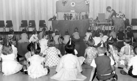 A group of children in party hats watch a puppet show.