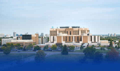 A large hospital complex with multiple buildings is shown, surrounded by trees and parking lots, with a cityscape visible in the background under a clear sky.
