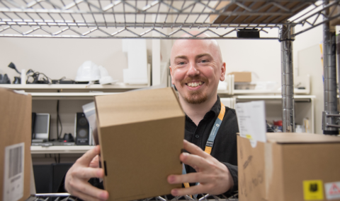 Team member smiles at camera while reaching for box of IT equipment from shelf
