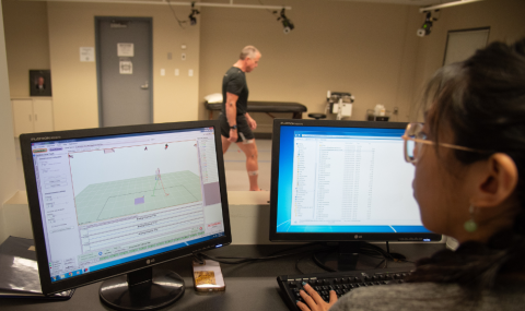 A person sits at a desk with two monitors, analyzing motion data, while another person stands in the background of a clinical setting.