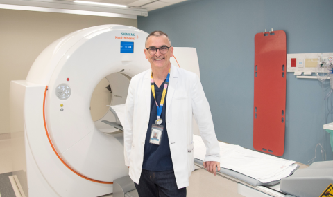 A doctor in a white coat stands in front of a PET scanner in a medical imaging room.