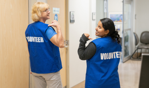 Two people wearing blue vests labeled "Volunteer" stand in a hallway, looking over their shoulders and pointing to the labels on their backs.