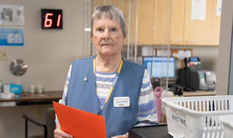 A woman wearing a volunteer vest holds an orange folder in a hospital clinic reception area, with office equipment and a digital clock displaying "61" in the background.
