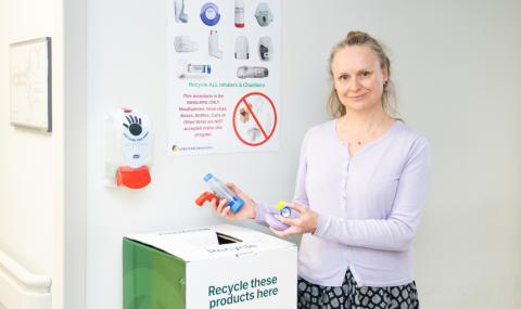 A woman stands by a recycling bin, holding inhalers, with a recycling poster for inhalers and a hand sanitizer dispenser on the wall behind her.