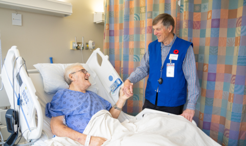 An elderly hospital patient in a hospital bed smiles and shakes hands with a volunteer in a blue vest standing by the bed.