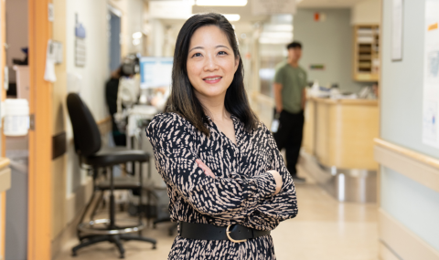 a woman stands with her arms crossed while smiling at the camera in a busy hospital hallways