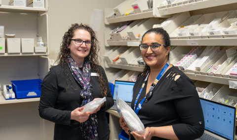 Female pharmacist holding pharmaceutical supplies smiles at camera standing next to female pharmacy resident who is also holding pharmaceutical supplies in a medication room