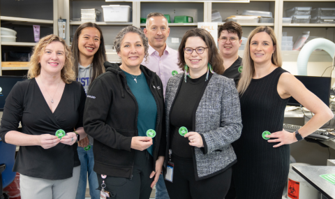 Seven people standing together in an office or lab setting, each holding a green circular badge and smiling at the camera.